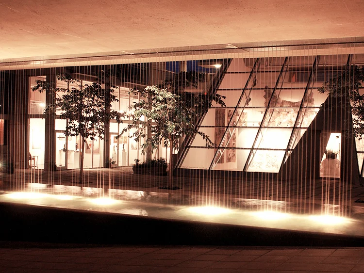 Vertical curtain of water at night, lit from above, with the glowing wedge-shaped glass restaurant pavilion visible through the water.